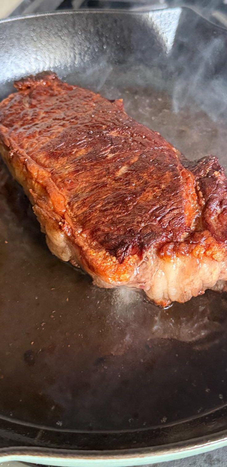 New York strip steak searing in a cast iron pan, deep mahogany crust.